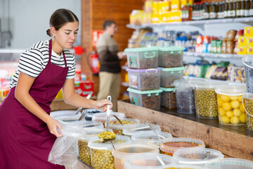 Seller girl in supermarket scooping marinated green olives