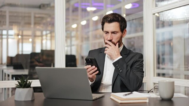 Male employee businessman sits at his desk in the office, looking concerned as he reads shocked bad message on phone. Executive manager has a laptop, notepad, pen, and coffee cup on his desk.