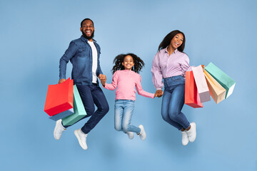 Sales And Discounts. Full body length shot of excited African American family of three people jumping, holding hands and shopping bags with new clothes, rejoice purchase, blue studio wall