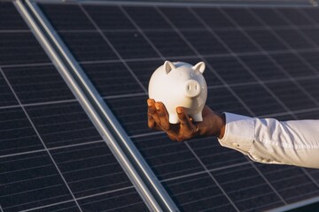 Businessman Holding Piggy Bank Over Solar Panels for Eco Investment © anatoliycherkas