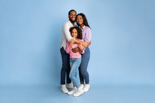 Happy Loving Family. Vertical full body length shot of African American woman and man embracing smiling girl standing isolated on blue studio wall. Cheerful mother and father posing with daughter