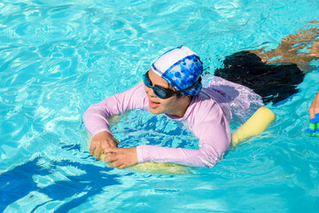 Happy girl with Down syndrome enjoying summer pool fun, wearing swim goggles and protective shirt...