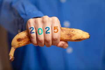 Man hand in shirt holds banana. Numbers 2025 on fingers