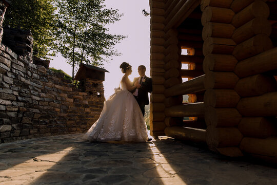A bride and groom stand in front of a log cabin, with the bride wearing a white dress