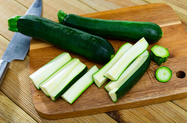 Against background of wooden table, several large pieces of zucchini are laid out on board. Ripe vegetables on chopping board before cooking dinner. Cut some zucchini fruit into oblong segments, parts