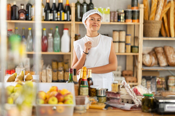 In sales area of store, young woman seller is waiting for visitors. Worker in apron uniform sells beer and wine, shows wide range of alcoholic beverages, offer meat delicious, vacuumed snacks.