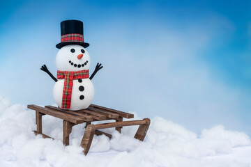 A happy snowman in a top hat and scarf, arms up and ready for winter fun! Posing with a sled on a snowy background and bright blue sky.