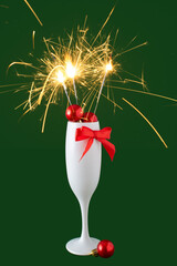 Champagne glass full of christmas decoration, red bow with sparkler on green background. Celebration idea, Christmas or Happy New Year concept.