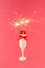 A champagne glass full of Christmas decorations, red bow with sparklers on pink background.  Christmas celebration or New Year party concept