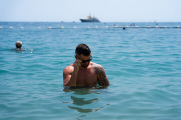 A man of European appearance, athletic build and wearing sunglasses swims on a beach in Monaco on a summer day.