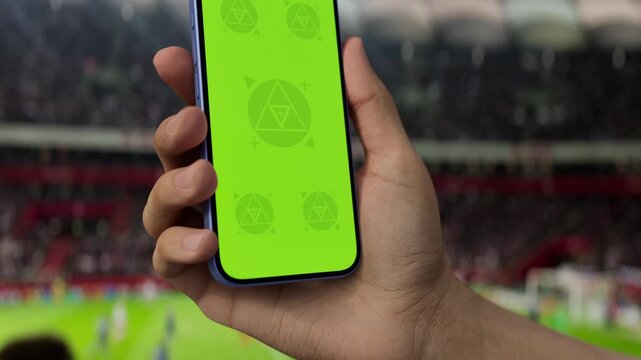 GREEN SCREEN CHROMA KEY A hand holding a phone with a green screen at a stadium during a football match. Ideal mockup for sports, betting, or event apps