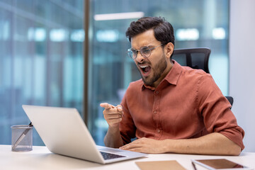 Business man showing frustration while pointing at laptop in office. Expresses emotions like anger or dismay during work. Seated at desk with stationery in modern environment.