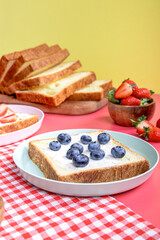 A slice of sandwich bread topped with cream and fresh blueberries on a plate. Background features more bread slices and a bowl of strawberries, creating a bright, fresh setup.