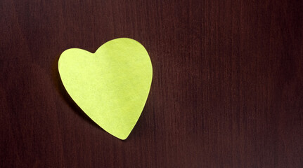 Fototapeta premium Yellow heart-shaped sticky note placed against a dark wood surface. The vibrant color contrasts with the background. Creating a simple yet striking visual ideal for concepts of love and positivity.