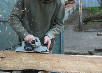 carpenter with electric plane sanding surface of wooden material in outdoor workshop with flying sawdust, planing texture of solid wood with hand tool