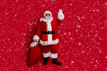 Full body photo of funny santa wave hold bag with presents wear hat eyewear coat pants boots isolated on red background