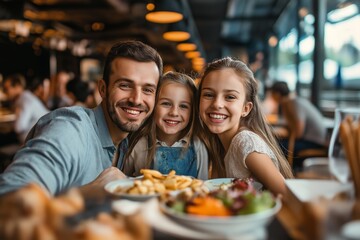 A man and two children are smiling and posing for a picture at a restaurant. The man is wearing a blue shirt and the children are wearing white shirts. The table is filled with food