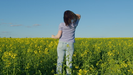 Fototapeta premium Woman In A T-Shirt Standing In A Flowering Canola Rapeseed Field. Pretty Young Woman With Arms Raised In Air In Rapeseed Field.