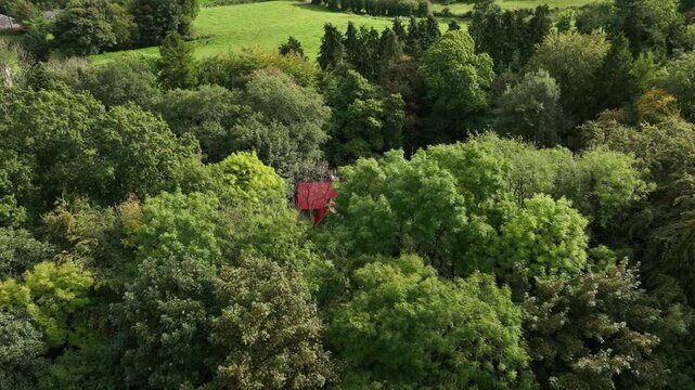 Saint Peter's Tin Church, Laragh, County Monaghan, Ireland, September 2022. Drone orbits clockwise above the vibrant trees revealing the red roof of the Swiss Gothic style building on a sunny day.