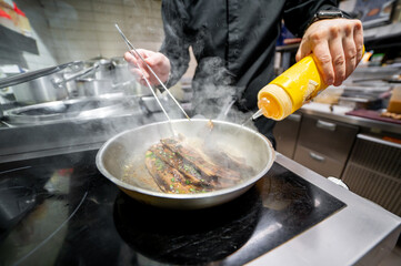 A chef in a professional kitchen adds sauce to a sizzling pan with food, steam rising from the hot pan. The scene captures the dynamic and vibrant atmosphere of culinary preparation.