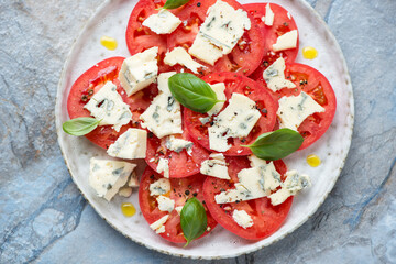 Plate with sliced red tomatoes, blue cheese and green basil, horizontal shot on a blue stone background, high angle view