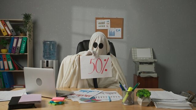 Halloween businessman ghost in white sheet sits at office desk holds shows paper sign with 50 percent discount text at camera. Happy halloween business concept.