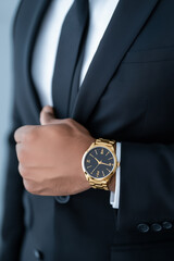 A man is adjusting his tie, elegantly dressed in a black suit, highlighting his fashionable gold wristwatch