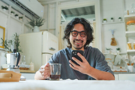 Young Asian man with eyeglasses and beard smiling while using mobile phone and drinking coffee in a kitchen. Japanese male relaxing with smartphone at home.