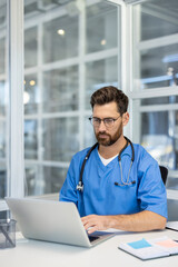 Fototapeta premium Medical professional wearing blue scrubs and glasses working on laptop in bright modern office. Represents healthcare technology, telemedicine, healthcare administration. Office environment suggests