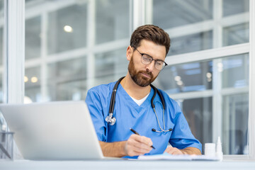 Doctor in blue scrubs writing at desk with laptop in modern office. He is focused, wearing glasses...