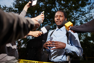 Low angle portrait of young African American man as police officer talking to reporters on crime...
