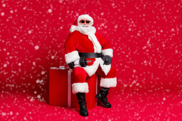 Full length photo of retired old man white beard sit big giftbox hold dumbbells warming up arms wear x-mas santa costume glove coat belt sunglass cap boot isolated red color background