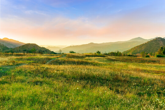 rustic beautiful lendscape of sunset field with green reeds and golden grass and amazing mountain valley with bright cloudy evening sky on background