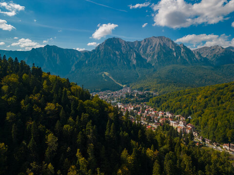 A town at the base of the mountain in the autumn season. Astonishing aerial view with the rocky mountains filmed in cinematic style. Romanian landscape above Busteni city in a sunny day with blue sky