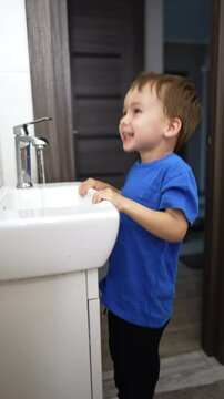 Happy smiling baby holds by the sink and jumps joyfully. Active toddler looks at running water and closes the faucet. Vertical video.
