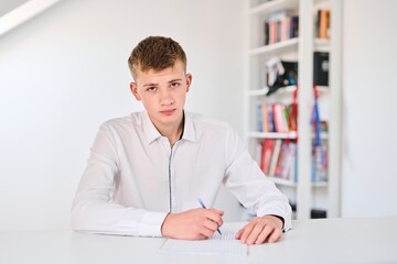 Fototapeta premium Portrait of a high school student in a white uniform doing homework, sitting at a table with textbooks, writing in a notebook