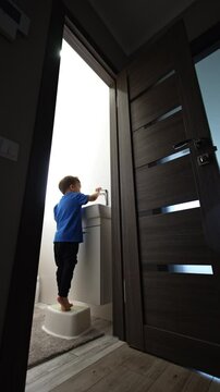 Toddler kid enters the bathroom and stands on the chair near the sink. Baby boy plays with water and jumps happily. Low angle view. Vertical video.
