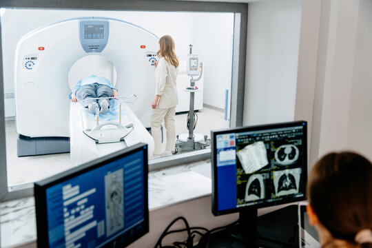 Caucasian male patient undergoing CT scan procedure while female technician supervises. Medical professionals examining diagnostic images on monitors in hospital setting, technology and healthcare.