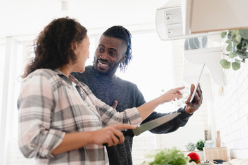 African-american husband helping his wife cook breakfast dinner at home kitchen showing digital...