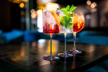 Three colorful cocktails with ice and garnishes on a bar counter, with a blurred background....