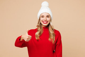 Merry fun young woman wear red warm cozy knitted sweater hat posing showing thumb up like gesture isolated on plain pastel light beige background. Happy New Year celebration Christmas holiday concept.