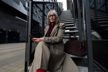 A middle-aged businesswoman seated on stairs, checking her phone outside a modern building on a...