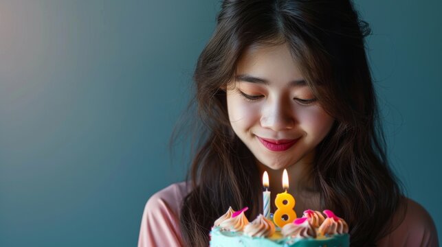 Joyful young woman celebrating her 18th birthday with a colorful cake featuring a lit number 18 candle. She smiles warmly, enjoying the special moment in front of a blue background.