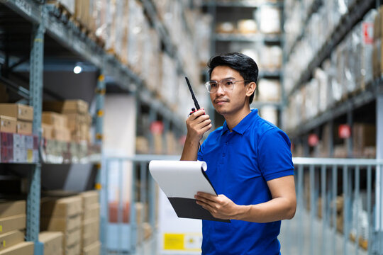 A man in a blue shirt is standing in a warehouse with a clipboard in his hand. He is wearing a headset and he is talking on the phone. Concept of work and productivity