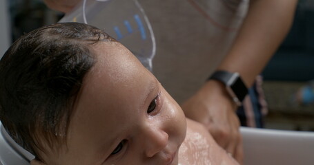 Water gently pouring over baby’s body from a cup during bath, baby attentive and calm, focus on...
