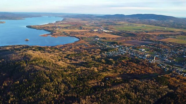 High-altitude aerial drone shot unveiling the village of Sayabec along Lake Matapedia, surrounded by early cold-season foliage and muted yellow tones in the scenic Matapedia Valley, Quebec