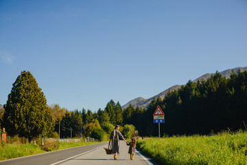 Obraz premium Mom and daughter walk along the road by the hand against the background of the forest and mountains. Mom and a girl in dresses. There is a picnic basket in his hands. Italian alps, countryside, tranqu