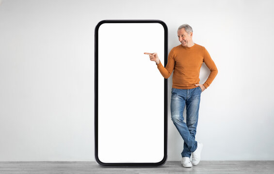 Fototapeta A man in a brown sweater and jeans stands next to a large, empty smartphone display, posing with a playful gesture. The setting is bright and minimalistic, emphasizing the display.