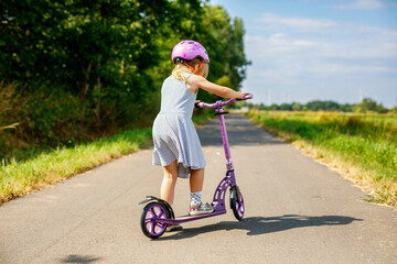 Portrait of active little preschool girl with helmet riding scooter on road in park outdoors on summer day. Seasonal child activity sport. Healthy childhood lifestyle