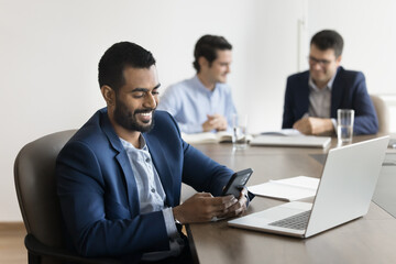 Happy Indian business project leader man typing on mobile phone at laptop, browsing social media, using Internet technology for job communication in meeting room shared with team
